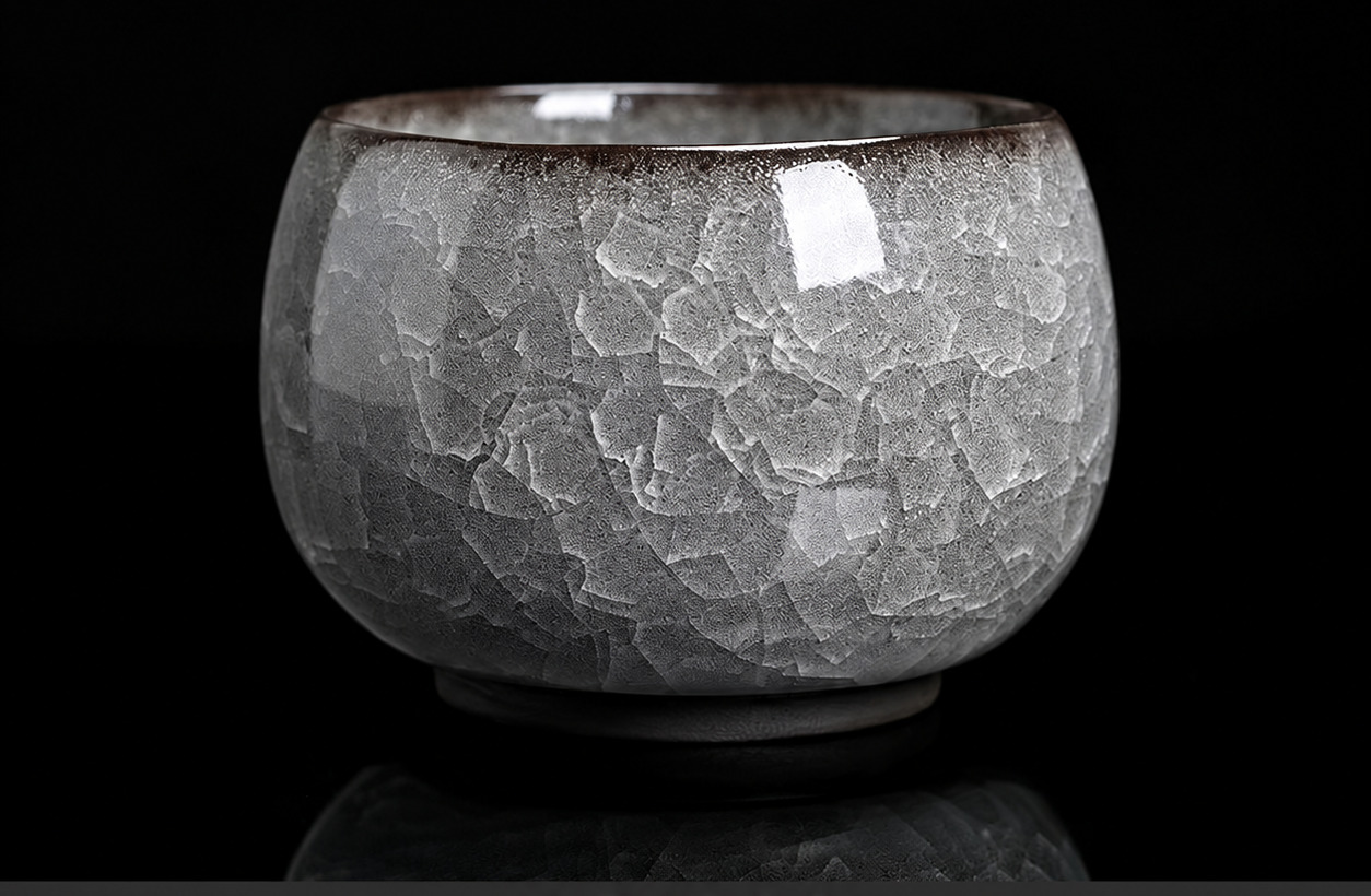 A rounded tea cup with a silver-grey ice-crackle crystalline glaze against a black background.