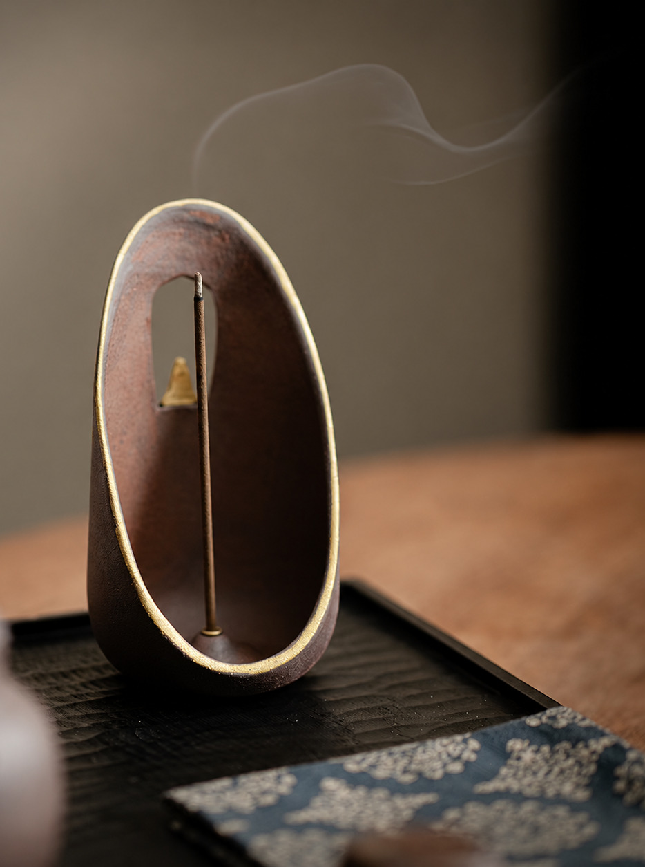 A dark oval incense holder with a brass rim holding a single incense stick, smoke rising.